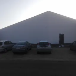 Budynek przemysłowy z płaską elewacją w trakcie prac wykończeniowych, widoczne zaparkowane samochody, instalacja klimatyzacji na ścianie, oraz osoba pracująca przy wejściu.