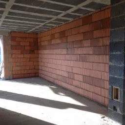 Wnętrze surowego domu w trakcie budowy, widoczne ściany z czerwonej cegły dziurawki, betonowy strop z pustaków oraz fragment muru z szarych bloczków betonowych z otworem wentylacyjnym, na podłodze...