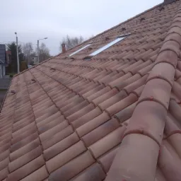 Dachówka ceramiczna w odcieniach brązu i czerwieni, z widocznymi dwoma oknami dachowymi i elementami mocującymi, sfotografowana pod kątem w pochmurny dzień.