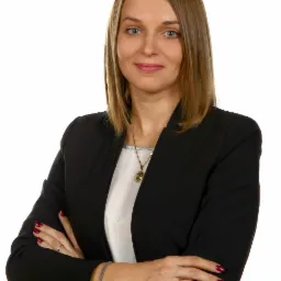 Portret kobiety w czarnej marynarce, z rękami skrzyżowanymi na piersiach, na białym tle. Widoczny delikatny uśmiech, biżuteria i staranny manicure.