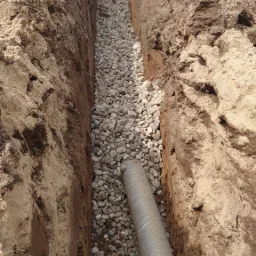 Wykop wypełniony drenażem z rurą perforowaną, widoczne fragmenty maszyny budowlanej i pomarańczowe spodnie robocze.