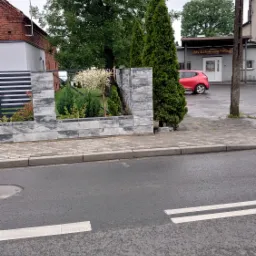 Nowoczesne ogrodzenie modułowe z szarego betonu w Lesznie, z widoczną roślinnością wewnątrz, w tle budynek i czerwony samochód.