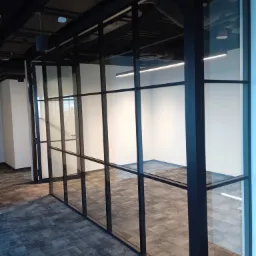 Szklana ścianka działowa z czarnymi profilami w biurze, widok na puste pomieszczenie z szarą wykładziną dywanową, oświetlenie sufitowe LED.