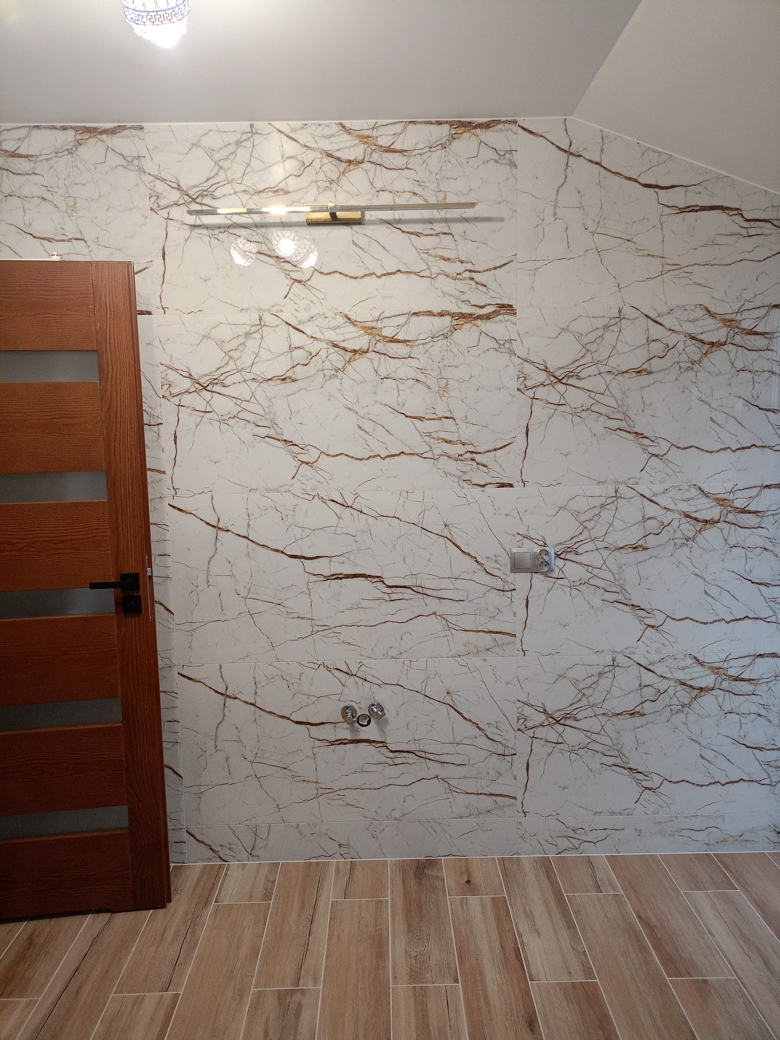 Wykończona łazienka z płytkami imitującymi marmur na ścianach, drewnopodobnymi płytkami na podłodze, drewnianymi drzwiami z przeszkleniem i oświetleniem punktowym.