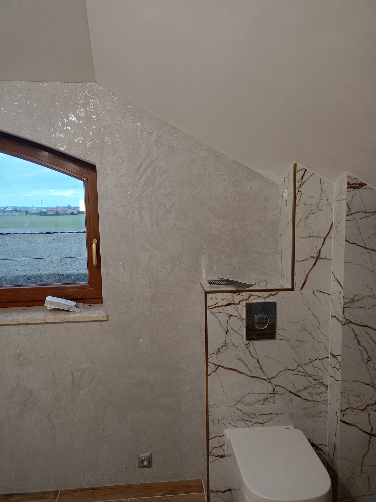 Wykończona łazienka na poddaszu ze ścianami pokrytymi dekoracyjnym tynkiem w odcieniach szarości z połyskującymi drobinkami, marmurowe płytki z brązowymi żyłami na ścianie z toaletą, okno dachowe...