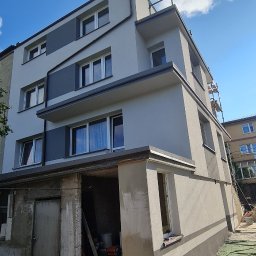 K . D INVEST Karol Damps Usługi ogólnobudowlane - Wielopiętrowy dom z szarą elewacją w trakcie prac wykończeniowych, widoczne worki z zaprawą budowlaną, rusztowania i fragment nieotynkowanej piwnicy.