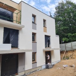 K . D INVEST Karol Damps Usługi ogólnobudowlane - Dwukondygnacyjny dom w trakcie budowy, częściowo ocieplony styropianem, z oknami zabezpieczonymi folią, malowany na biało, z widocznym rusztowaniem i pracownikiem malującym elewację przy wejściu.