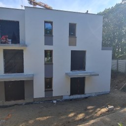 K . D INVEST Karol Damps Usługi ogólnobudowlane - Dwukondygnacyjny dom w trakcie budowy, z oknami zabezpieczonymi folią, pracownik siedzący na balkonie przy wiadrach z farbą, świeżo pomalowana elewacja w odcieniach bieli i szarości, widoczny żuraw...