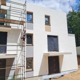 K . D INVEST Karol Damps Usługi ogólnobudowlane - Dwóch pracowników na rusztowaniu przy białym, dwupiętrowym domu w trakcie budowy, z oknami zabezpieczonymi folią i fragmentami elewacji w odcieniach szarości.