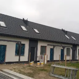 Szeregowy budynek w trakcie wykończenia elewacji, okna zabezpieczone folią, widoczne puszki z farbą i rusztowanie.