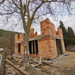 K . D INVEST Karol Damps Usługi ogólnobudowlane - Wznoszone ściany parterowego domu z czerwonej cegły ceramicznej porotherm, widoczne otwory okienne i drzwiowe, na pierwszym planie drzewo liściaste bez liści, w tle zieleń i zabudowa jednorodzinna.