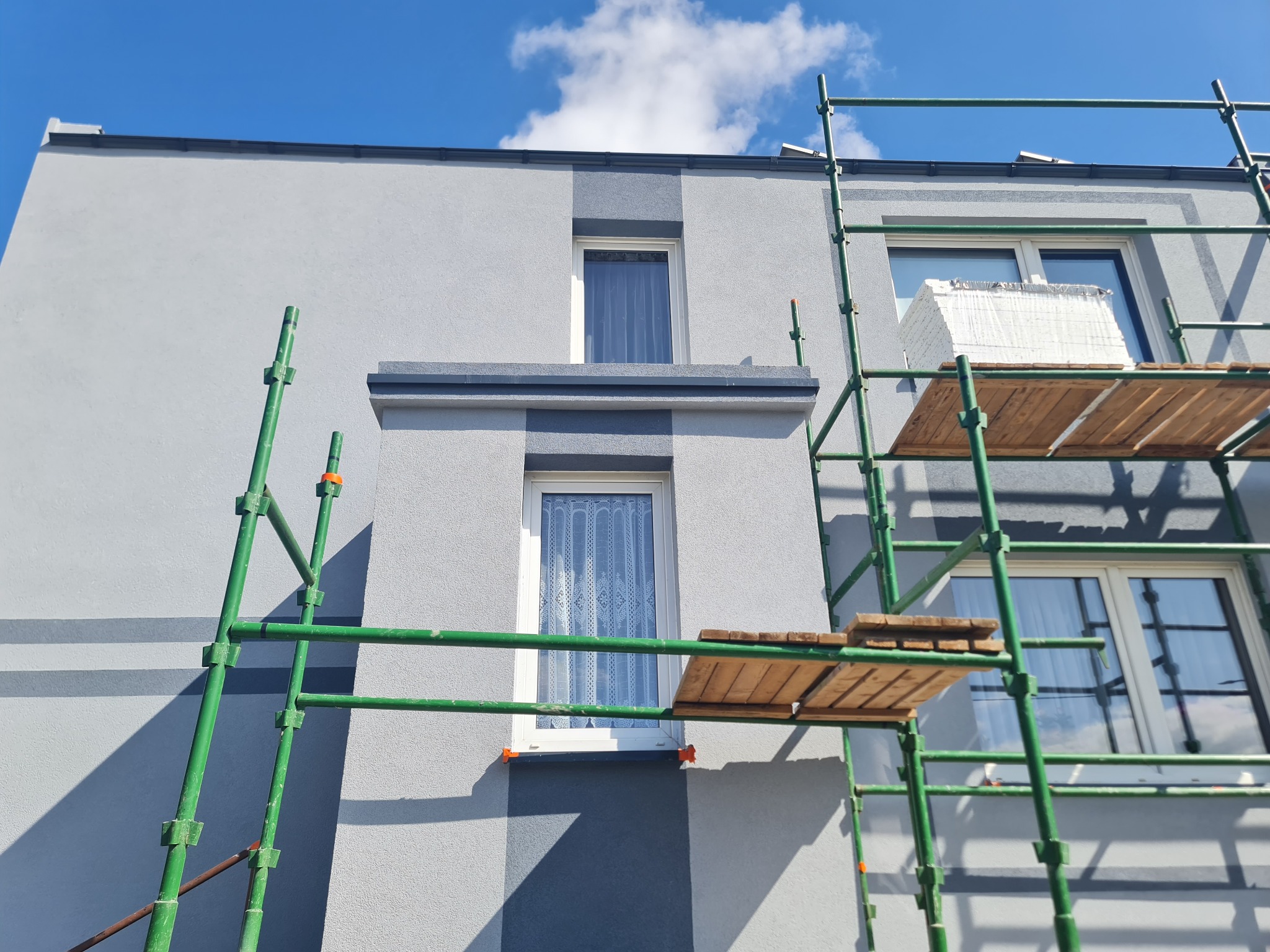 Szara elewacja domu w trakcie ocieplania, widoczne rusztowanie z zapasem styropianu na podeście, błękitne niebo w tle.