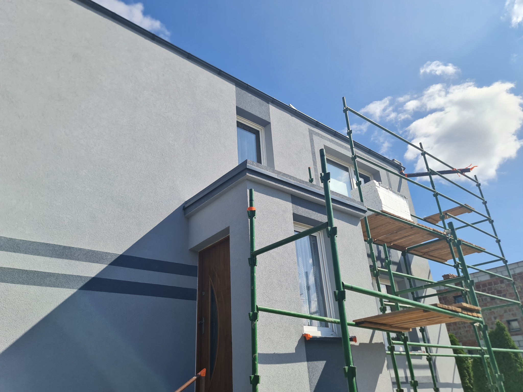 Szare, otynkowane ściany domu jednorodzinnego w trakcie prac wykończeniowych, z widocznym rusztowaniem i fragmentem nieba z chmurami.