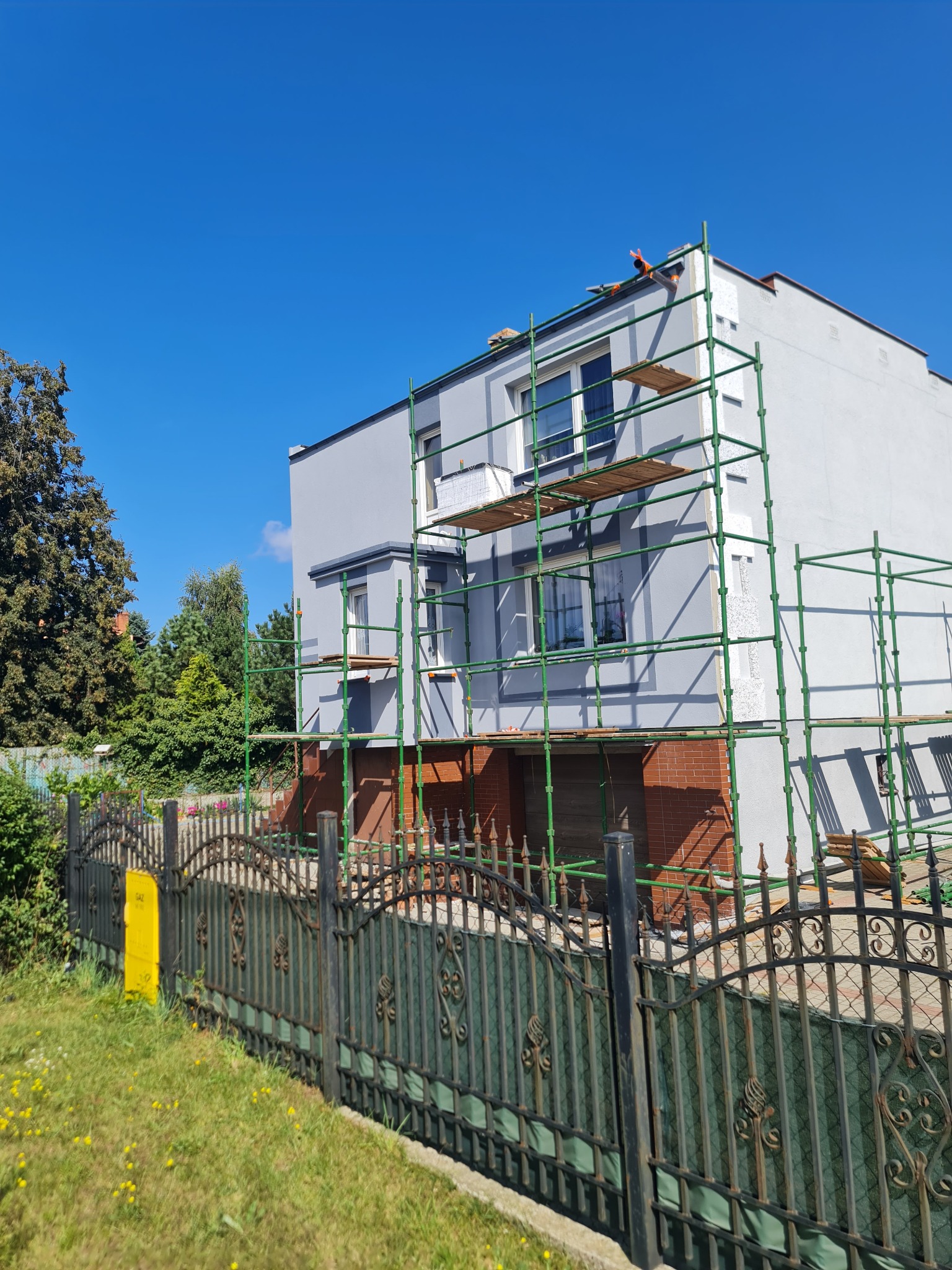 Dwukondygnacyjny dom w trakcie prac elewacyjnych, z zielonym rusztowaniem i częściowo pomalowanymi ścianami na szaro, widok zza metalowego ogrodzenia.