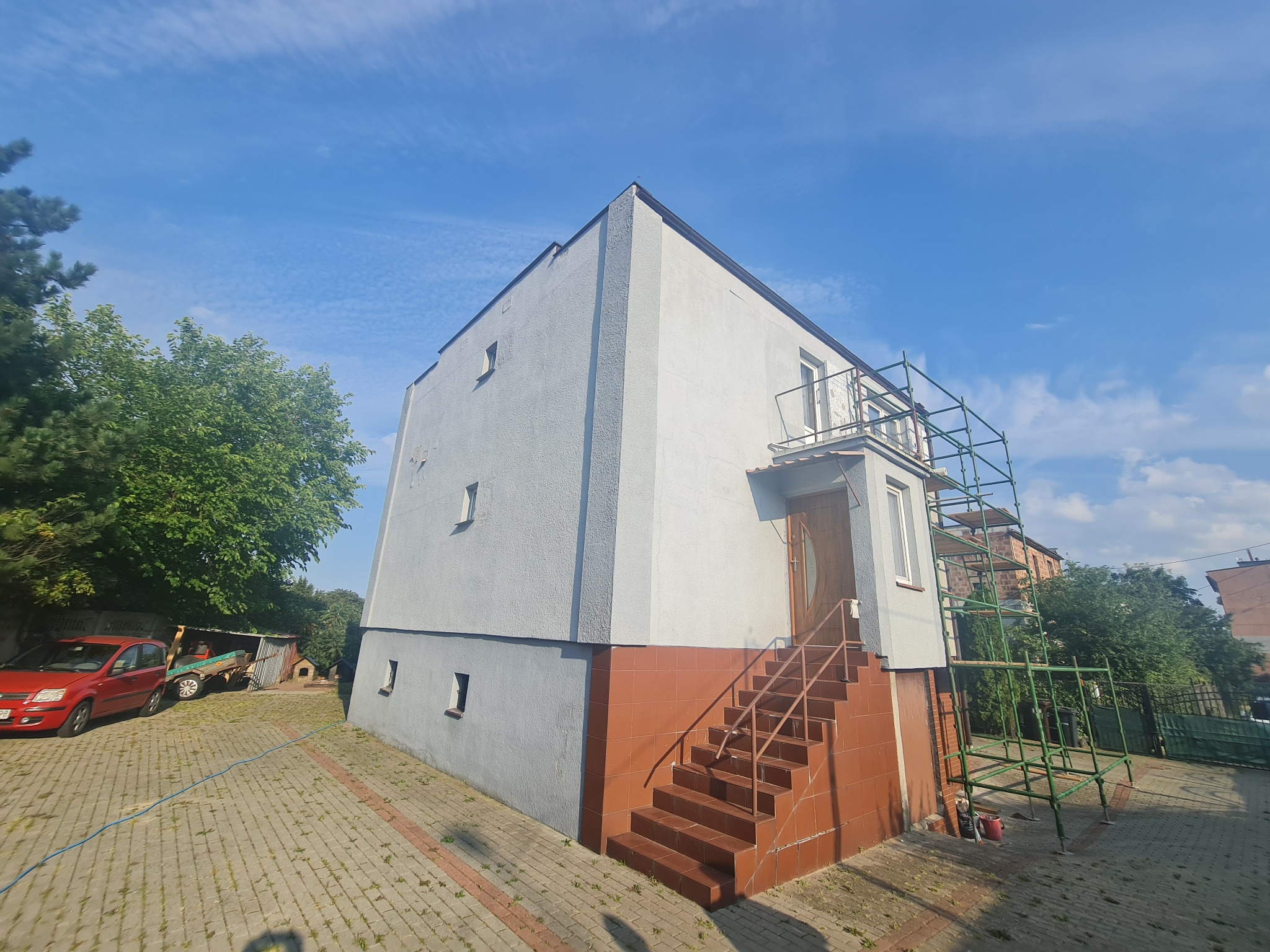 Dwukondygnacyjny dom z pomalowaną elewacją na szaro i biało, z brązowymi schodami wejściowymi, z zielonym rusztowaniem ustawionym przy balkonie, widziany z dołu pod kątem.