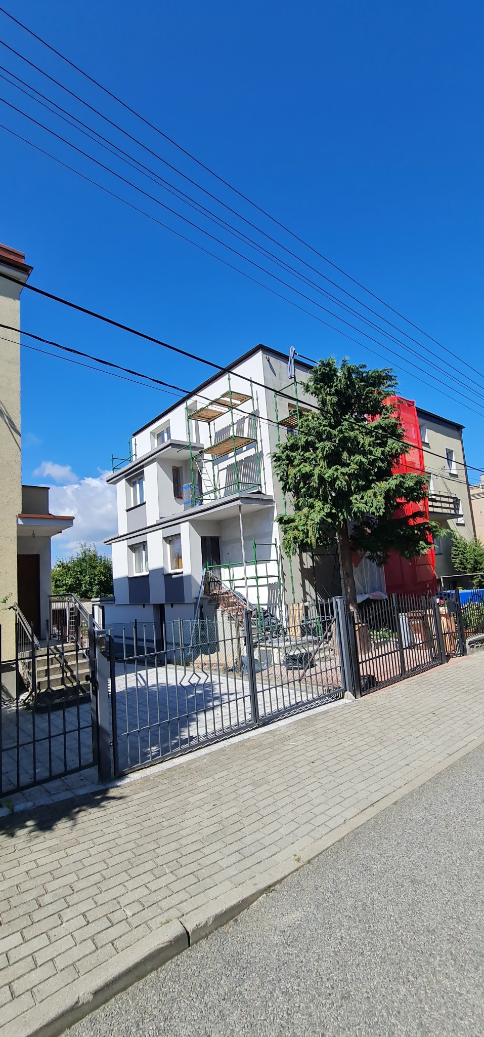 Dwupiętrowy dom w trakcie prac elewacyjnych, z widocznymi rusztowaniami i siatką ochronną, częściowo otynkowany na biało i szaro, z metalowym ogrodzeniem i drzewem w pierwszym planie.