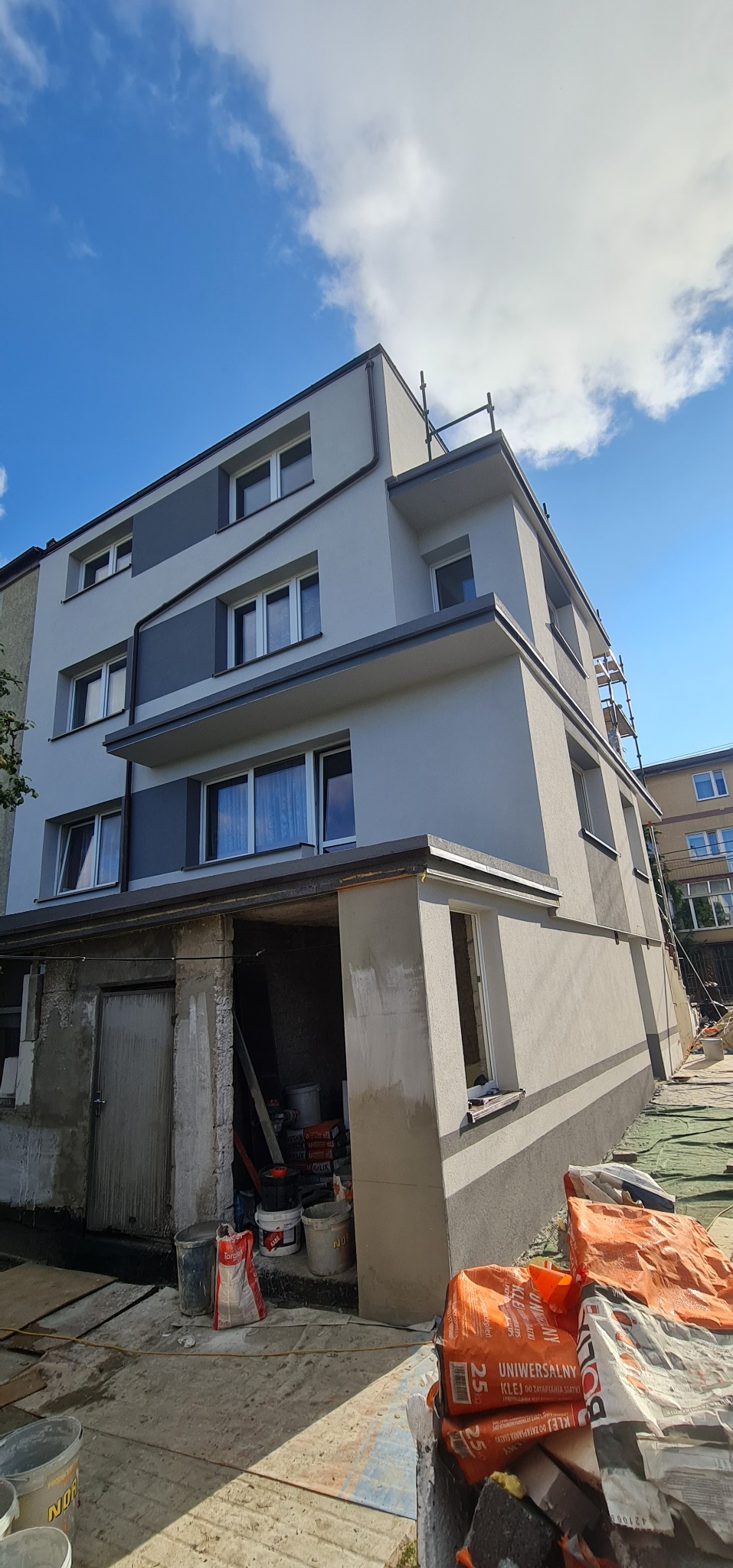 Wielopiętrowy dom z szarą elewacją w trakcie prac wykończeniowych, widoczne worki z zaprawą budowlaną, rusztowania i fragment nieotynkowanej piwnicy.