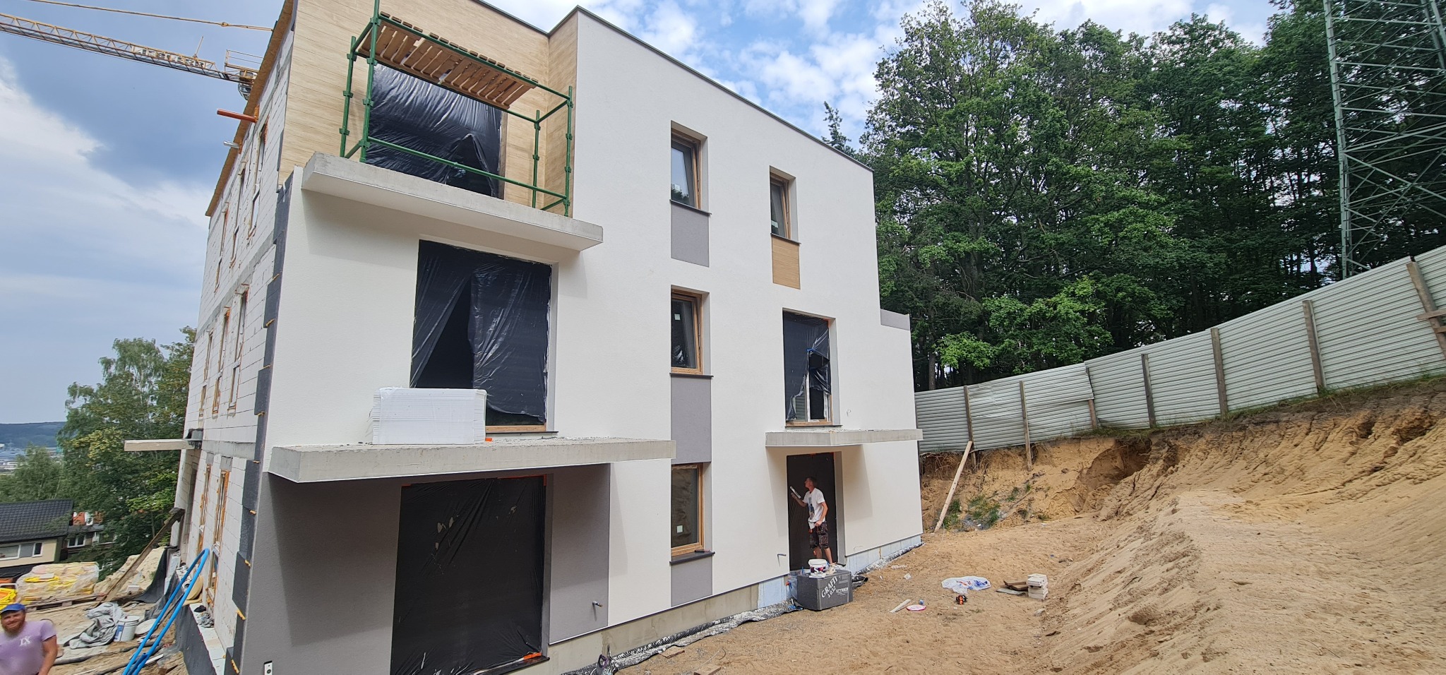 Dwukondygnacyjny dom w trakcie budowy, częściowo ocieplony styropianem, z oknami zabezpieczonymi folią, malowany na biało, z widocznym rusztowaniem i pracownikiem malującym elewację przy wejściu.