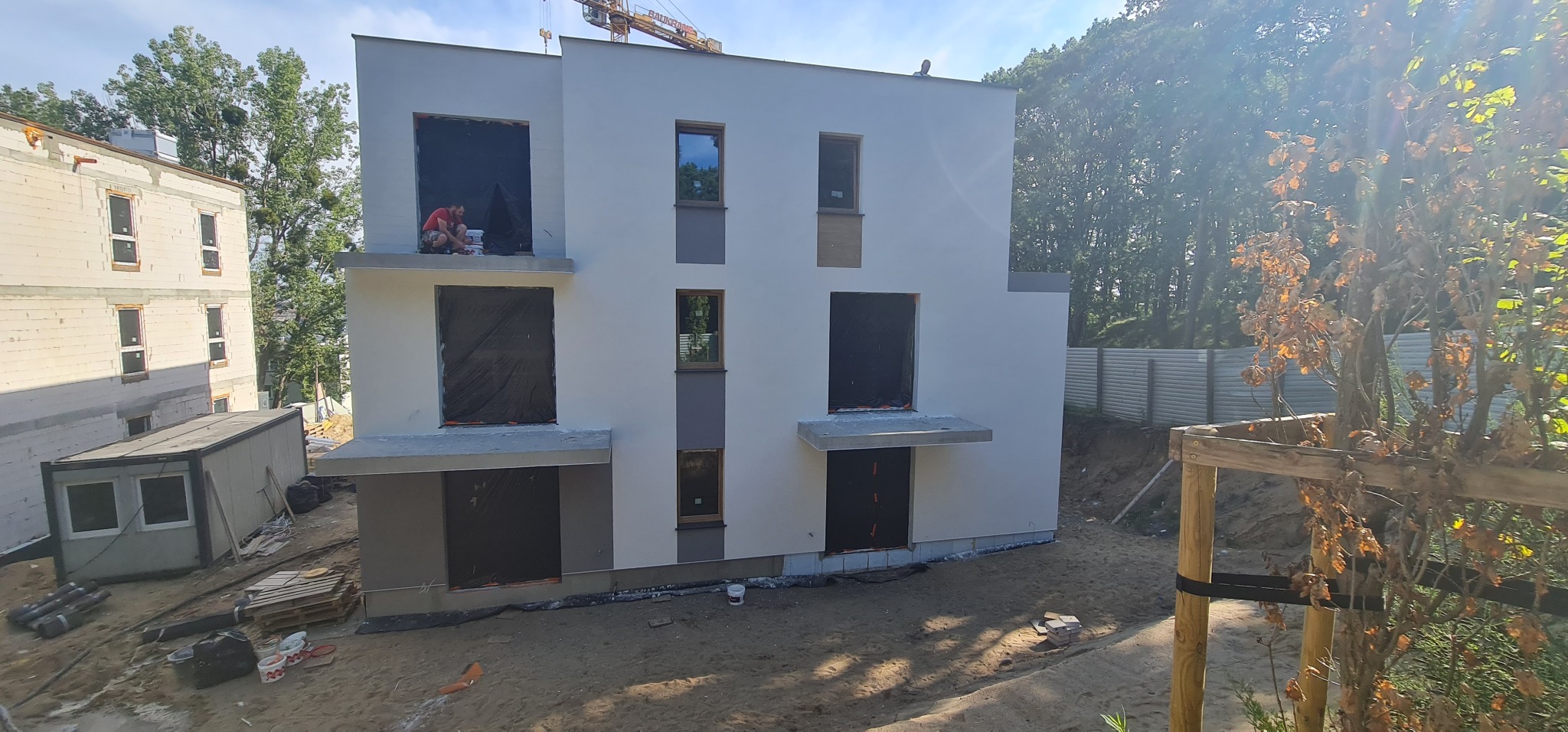 Dwukondygnacyjny dom w trakcie budowy, z oknami zabezpieczonymi folią, pracownik siedzący na balkonie przy wiadrach z farbą, świeżo pomalowana elewacja w odcieniach bieli i szarości, widoczny żuraw...