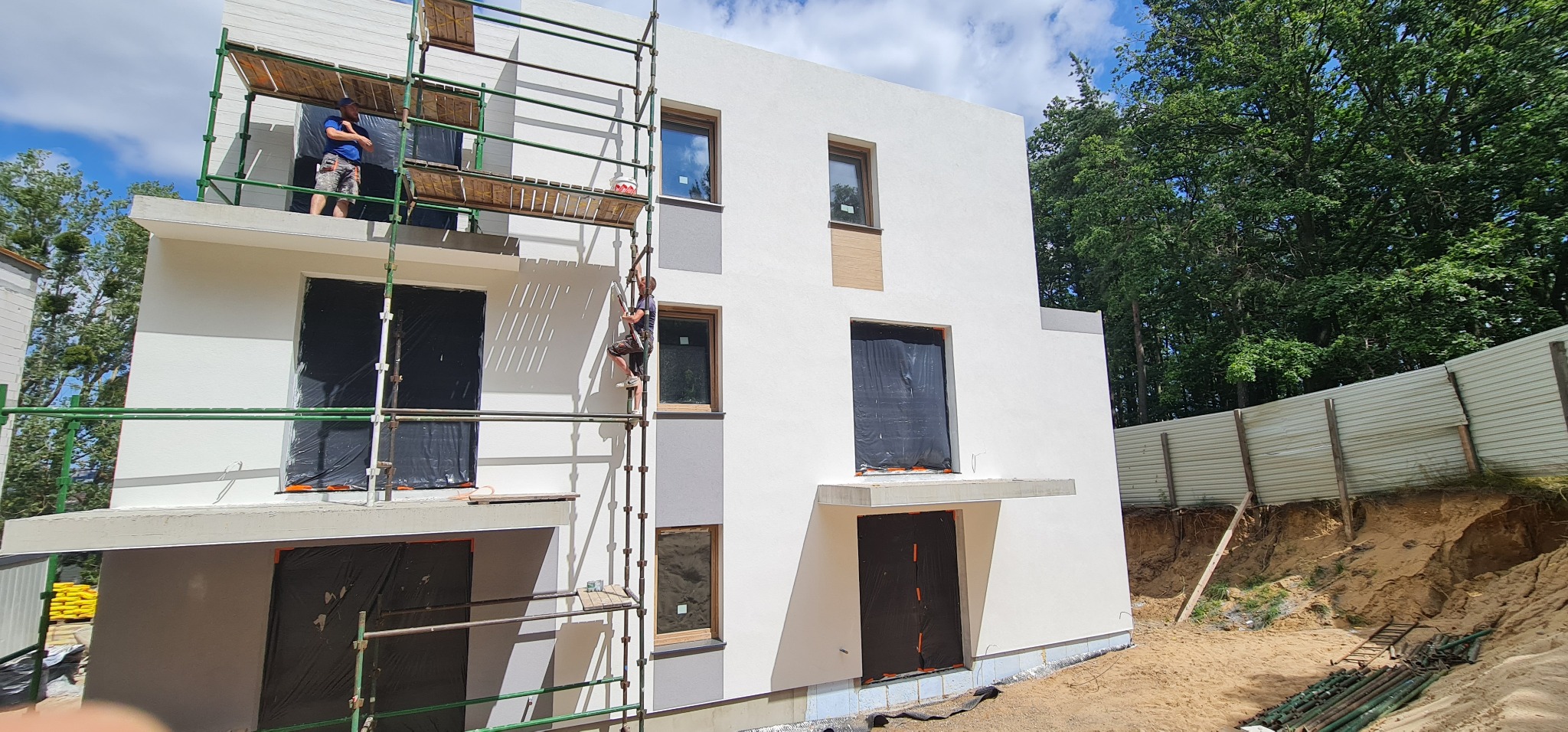 Dwóch pracowników na rusztowaniu przy białym, dwupiętrowym domu w trakcie budowy, z oknami zabezpieczonymi folią i fragmentami elewacji w odcieniach szarości.