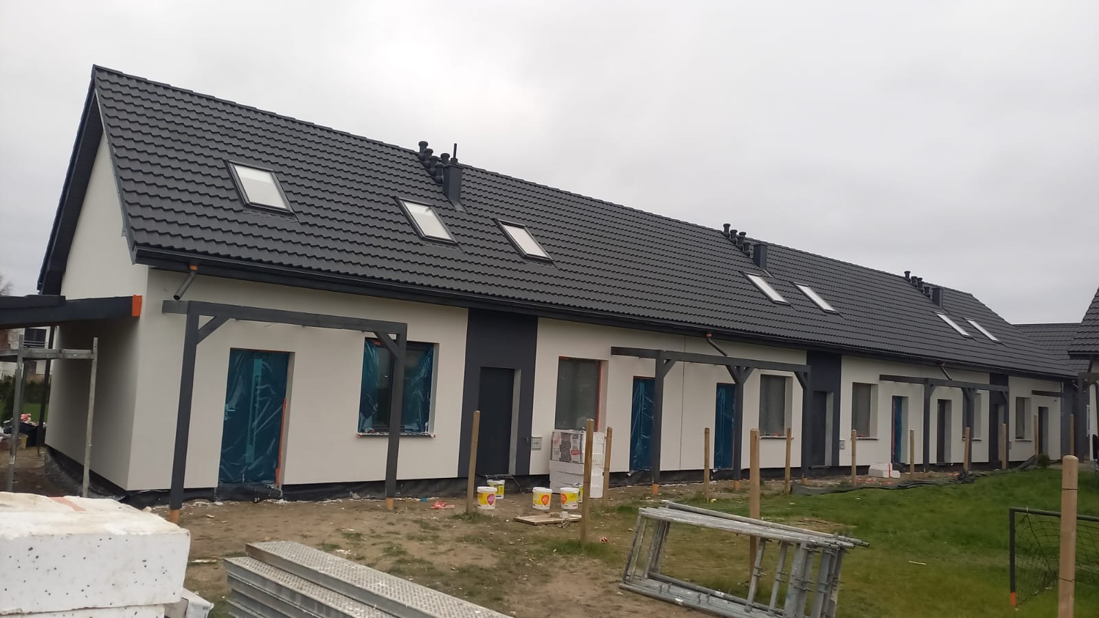 Szeregowy budynek w trakcie wykończenia elewacji, okna zabezpieczone folią, widoczne puszki z farbą i rusztowanie.