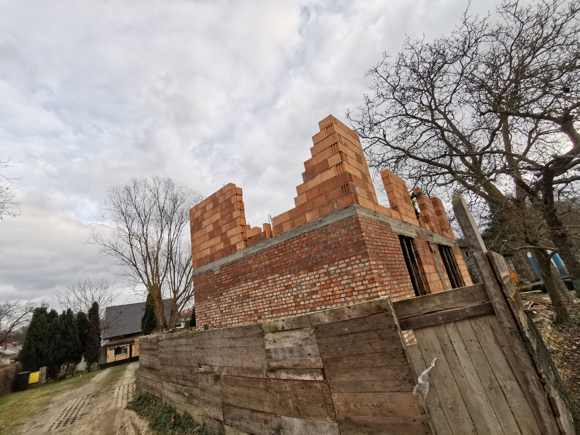 Wznoszenie ścian parterowego domu z czerwonej cegły i bloczków, widoczne drewniane ogrodzenie budowlane i pochmurne niebo.