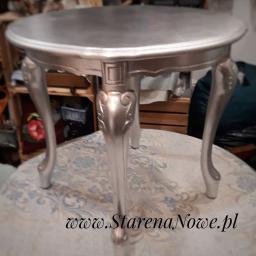 Renowacja Mebli Stare na Nowe - Okrągły, srebrny stolik kawowy w stylu retro z giętymi nogami, ustawiony na stole pokrytym tkaniną we wzory. W tle widoczne elementy wyposażenia wnętrza.