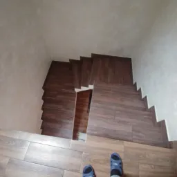 Widok z góry na schody wyłożone brązowymi płytkami imitującymi drewno, z fragmentem podłogi i stopami w niebieskich klapkach.