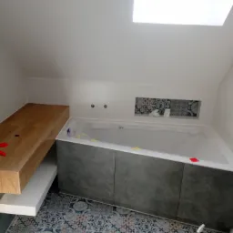 Wnętrze łazienki w trakcie prac: zabudowana wanna z szarą obudową, drewniany blat umywalkowy, wzorzysta glazura podłogowa i wnęka nad wanną, okno dachowe. Folia ochronna na wannie.