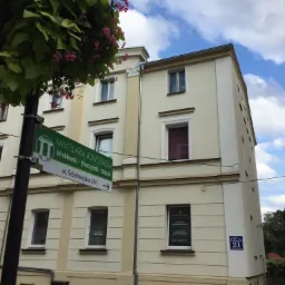 Zewnętrzny widok trzypiętrowego budynku z szyldem informującym o lokalizacji kancelarii adwokackiej, tabliczka z adresem 21, wisząca donica z kwiatami, niebieskie niebo z chmurami w tle.