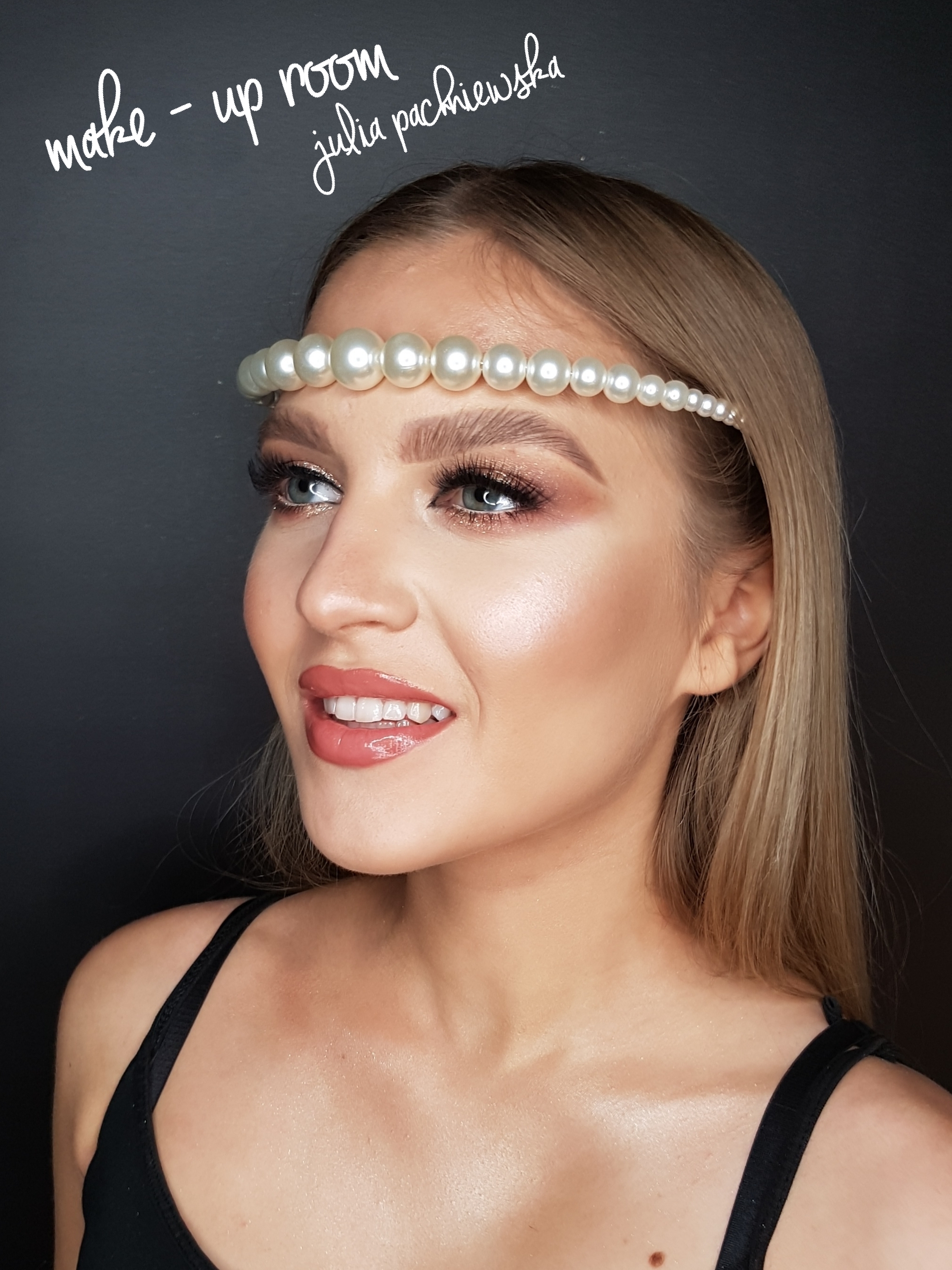 Portret uśmiechniętej blondynki z perłową opaską, w pełnym makijażu z błyszczącymi cieniami i błyszczykiem na ustach, na tle ciemnej ściany z napisem 'make-up room julia pachniewska'.
