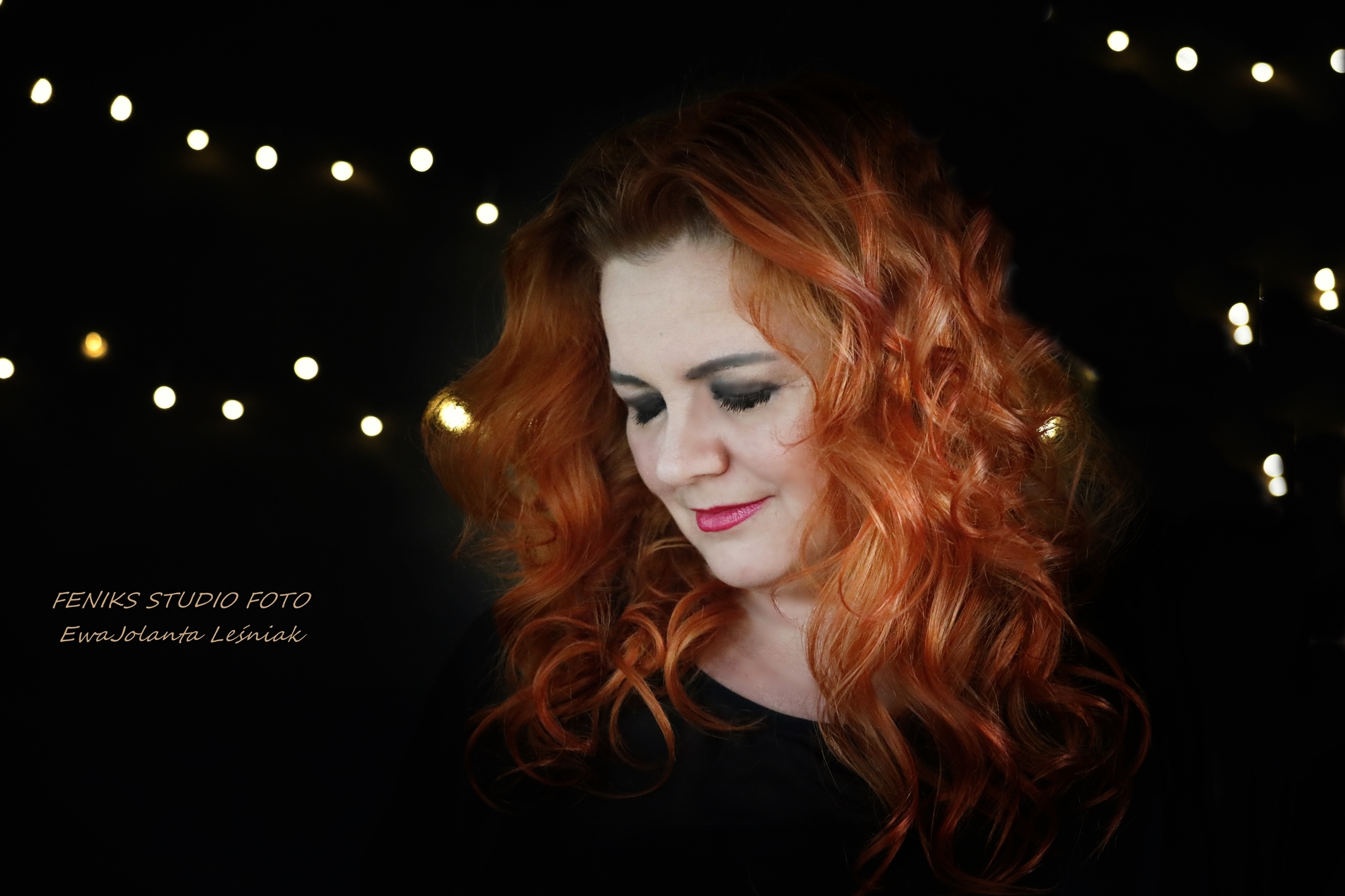Portret kobiety o rudych, kręconych włosach, z delikatnym uśmiechem i makijażem, na czarnym tle z rozmytymi światełkami.