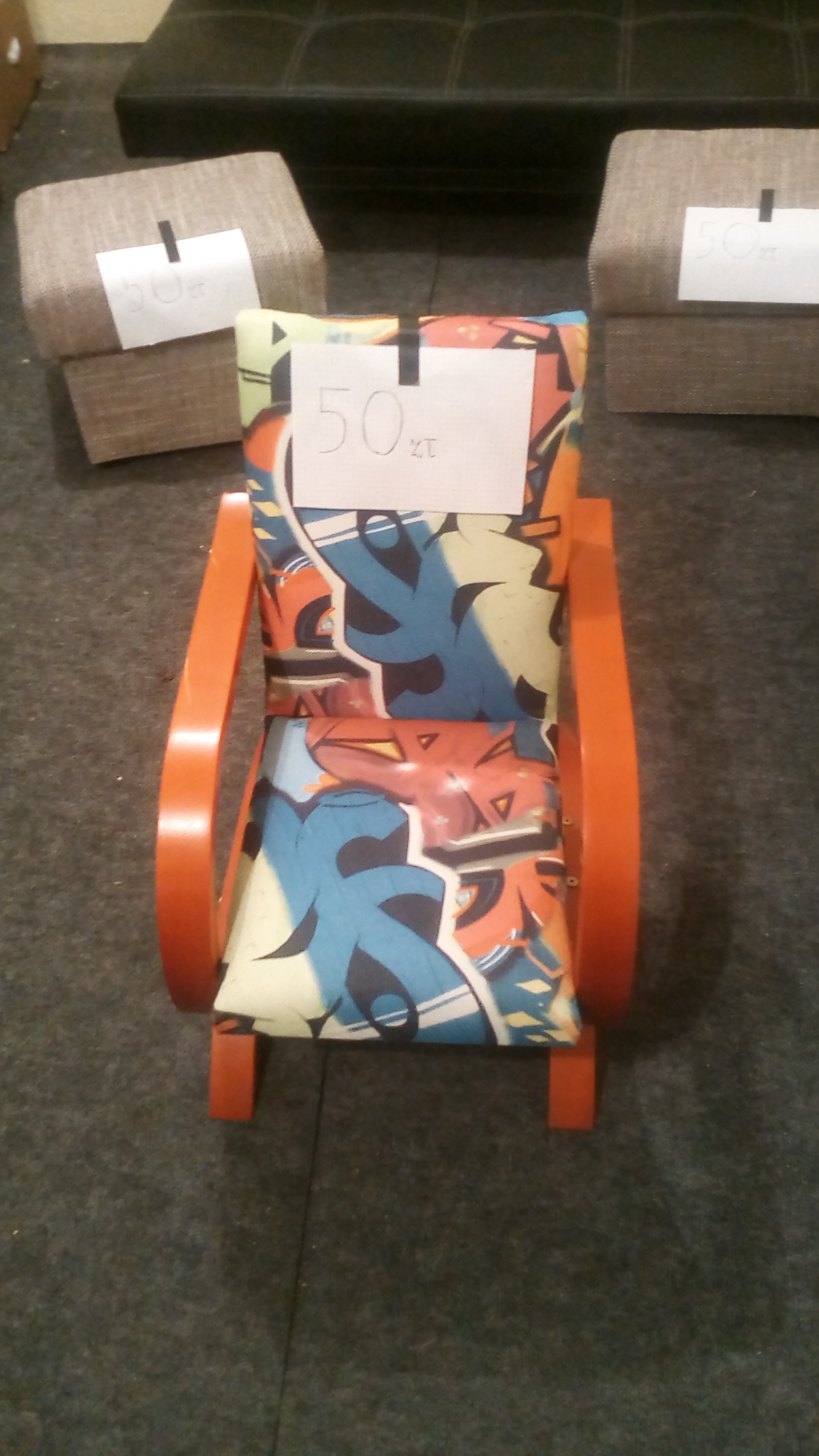 Mały, odnowiony fotel z pomarańczowymi drewnianymi elementami i tapicerką w graffiti, z ceną 50 zł, na tle dwóch taboretów i fragmentu sofy.