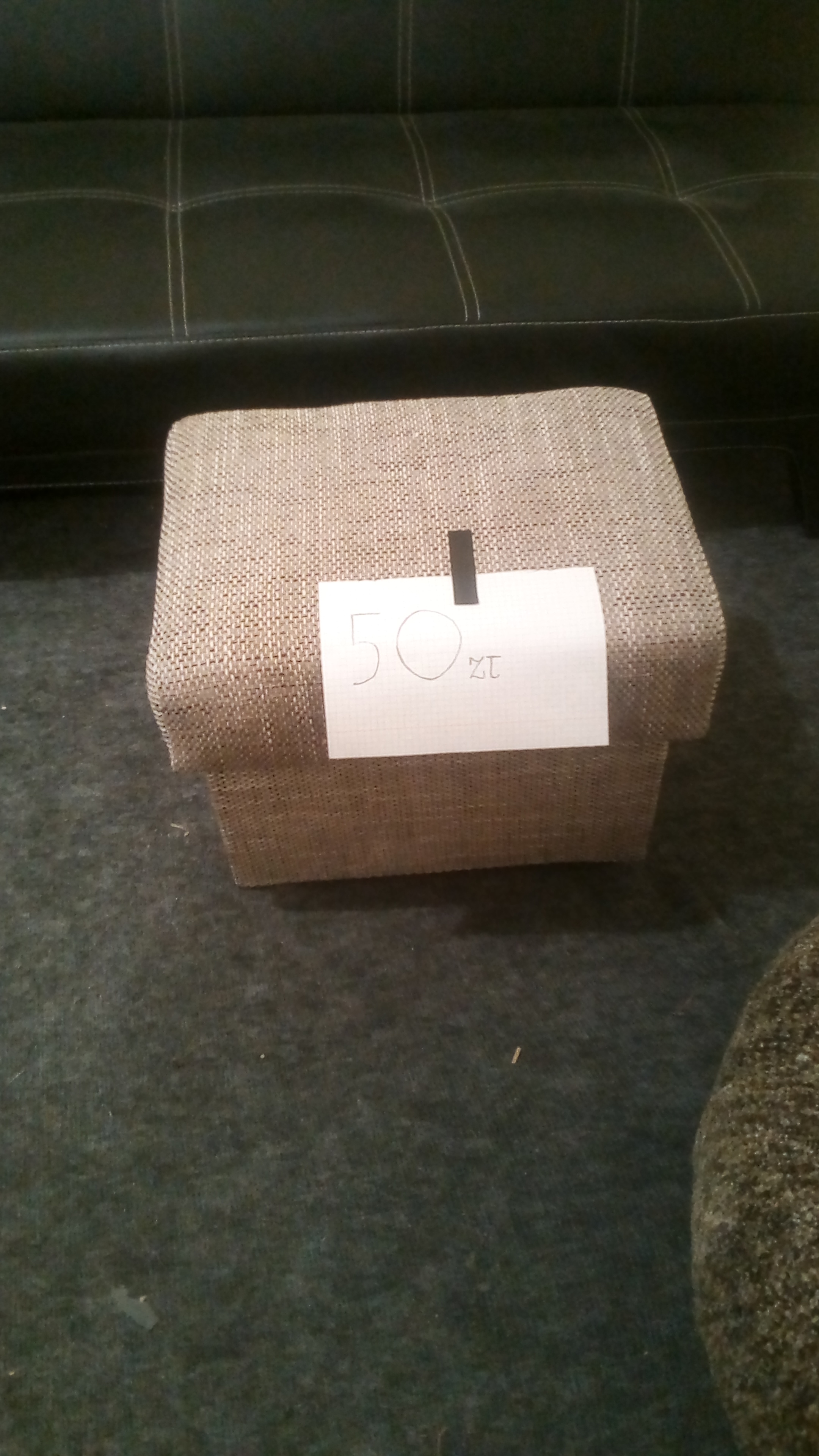 Beżowy, tapicerowany puf w kształcie sześcianu z kartką '50 zł' przypiętą spinaczem, stoi na ciemnozielonej wykładzinie dywanowej, z fragmentem czarnej sofy w tle.