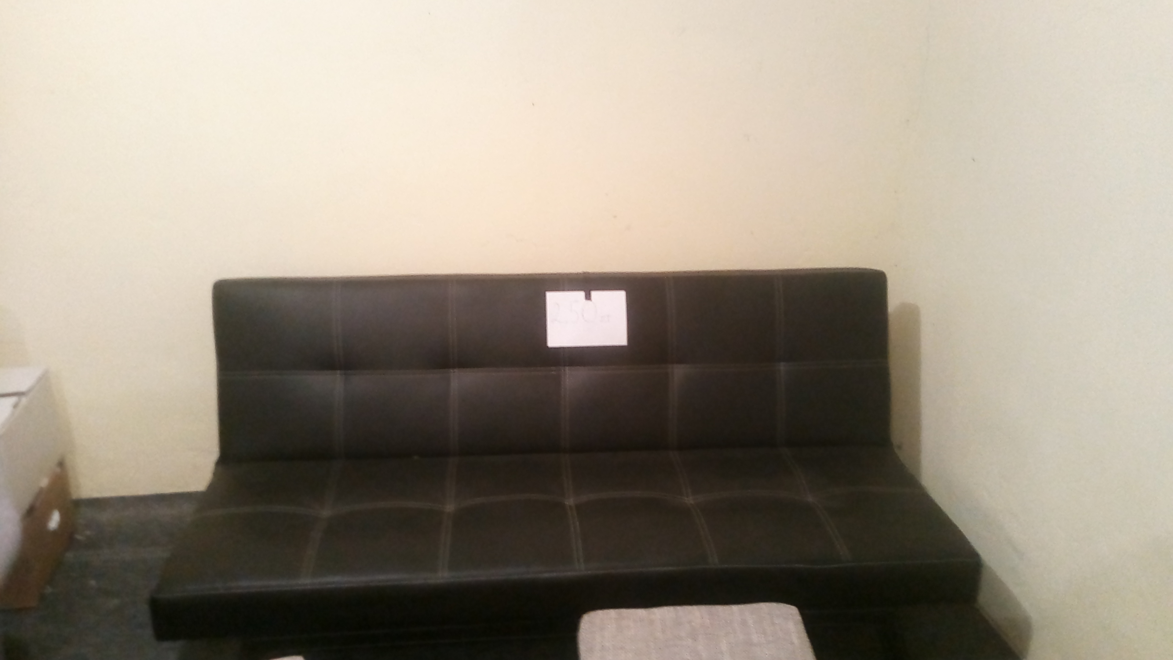 Ciemna, rozkładana sofa z kwadratowymi przeszyciami, częściowo widoczny tapicerowany podnóżek, na oparciu przyklejona kartka z napisem.