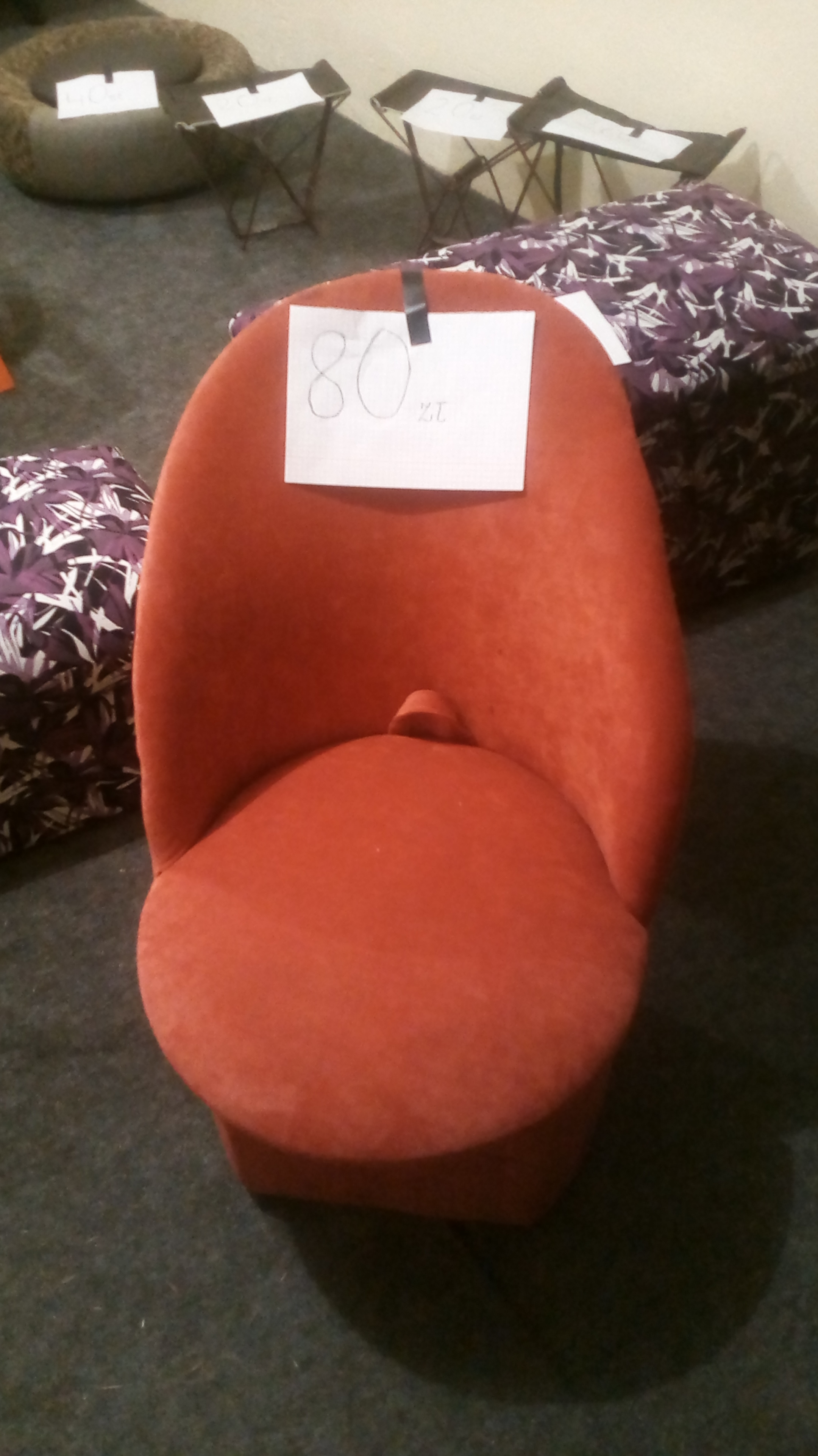 Pomarańczowe krzesło z ceną 80 zł na kartce, ustawione na szarym dywanie, w tle widoczne inne meble i akcesoria.