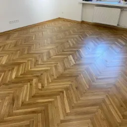 Wycyklinowany parkiet ułożony w jodełkę, widoczne ślady odbicia światła, ściany białe, grzejnik pod oknem.