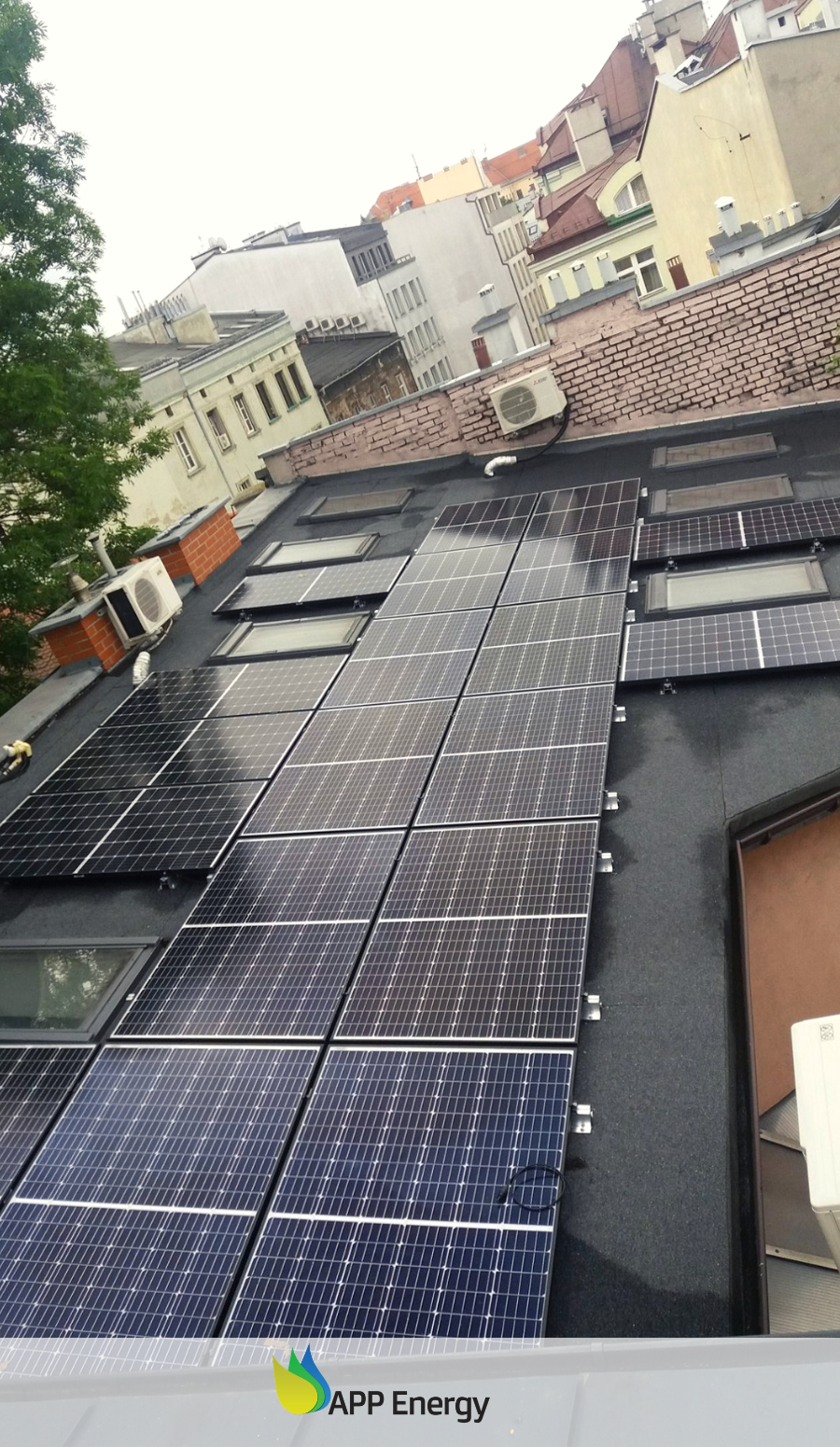 Instalacja fotowoltaiczna na płaskim dachu kamienicy z oknami dachowymi, widok z góry, pochmurna pogoda.