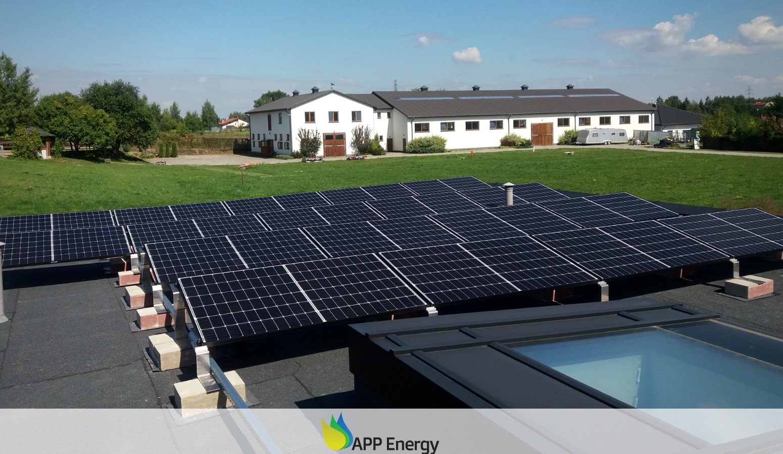 Instalacja paneli słonecznych na dachu budynku gospodarczego z widokiem na zielony trawnik i białe budynki w tle, widoczne ceglane podstawy pod konstrukcją montażową paneli.