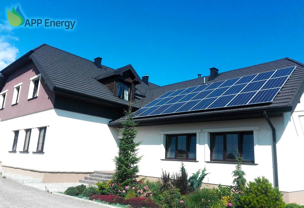 Dom jednorodzinny z ciemną dachówką i panelami słonecznymi na dachu, widok z ogrodu z ozdobnymi krzewami i drzewkami iglastymi. Logo firmy APP Energy w lewym górnym rogu.