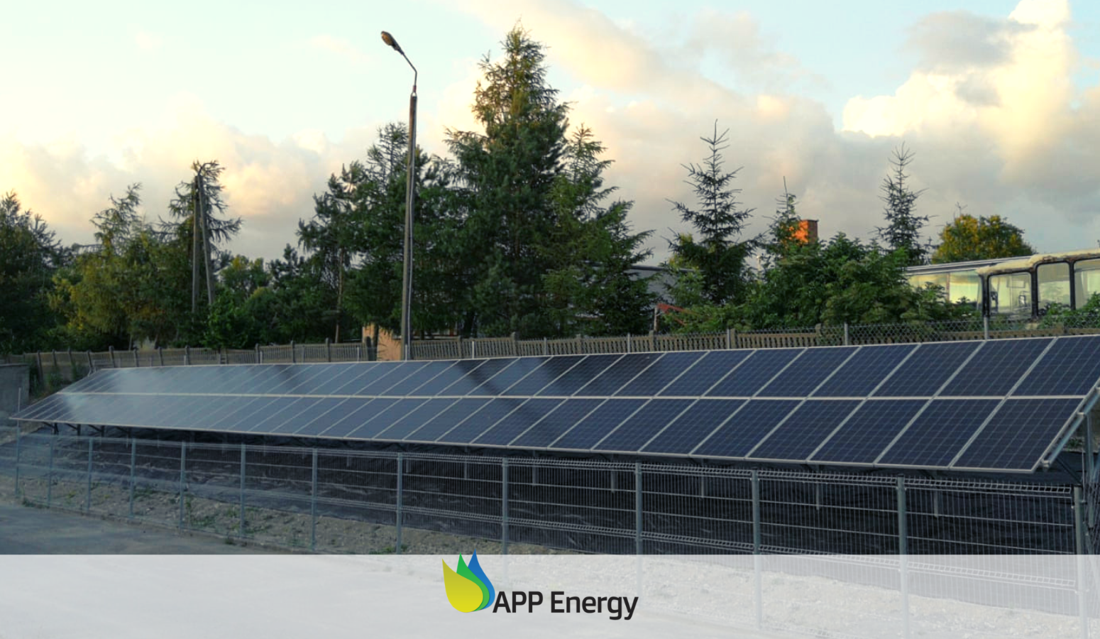 Długa instalacja paneli słonecznych za metalowym ogrodzeniem na tle drzew i zachmurzonego nieba o zmierzchu.