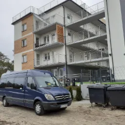 Niebieski bus Mercedes-Benz na piaszczystym podłożu przed nowoczesnym budynkiem apartamentowym z balkonami i numerem 4, obok czarne kontenery na śmieci.