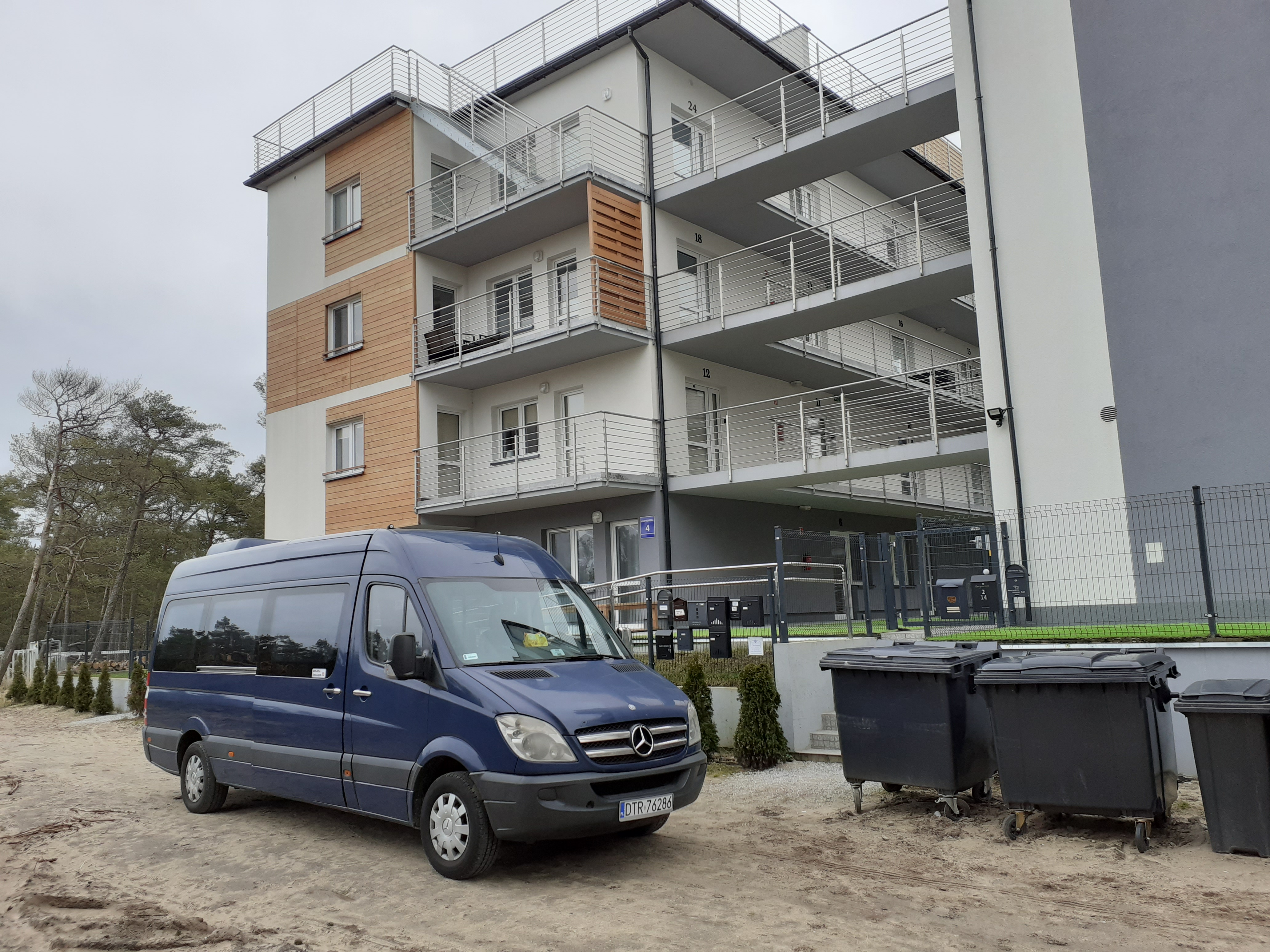 Niebieski bus Mercedes-Benz na piaszczystym podłożu przed nowoczesnym budynkiem apartamentowym z balkonami i numerem 4, obok czarne kontenery na śmieci.