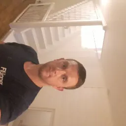 Mężczyzna w ciemnej koszulce z logo firmy malarskiej na tle świeżo pomalowanej, białej klatki schodowej z drewnianymi schodami.