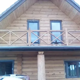 Dwukondygnacyjny budynek z bali z balkonem z drewnianą balustradą i ciemnymi oknami.