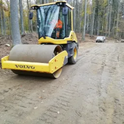 Żółty walec Volvo ubija grunt na leśnej drodze, w tle biały samochód dostawczy i drzewa.