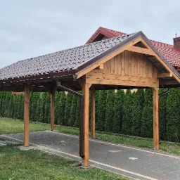 Drewniana wiata garażowa z brązowym dachem na posesji z tujami w tle, widok z boku.