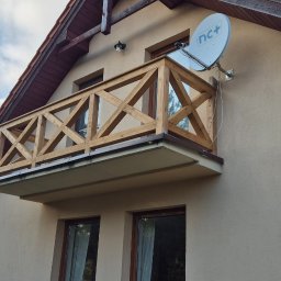 Drew-Tech Technikum Drewna - Balkon z drewnianą balustradą o geometrycznym wzorze krzyży, zamontowany na elewacji domu z brązową dachówką, widoczna antena satelitarna.