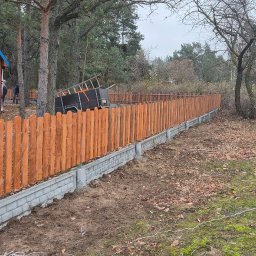 Drew-Tech Technikum Drewna - Nowo postawione, pomalowane drewniane ogrodzenie na betonowej podmurówce, widok na dom z niebieskim dachem i samochód dostawczy z narzędziami, teren częściowo pokryty mchem i liśćmi.