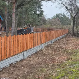 Nowo postawione, pomalowane drewniane ogrodzenie na betonowej podmurówce, widok na dom z niebieskim dachem i samochód dostawczy z narzędziami, teren częściowo pokryty mchem i liśćmi.